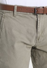 Chinos en coton kaki avec une texture légèrement froissée, dotés d'une ceinture en cuir marron et de détails de couture visibles au niveau de la taille.