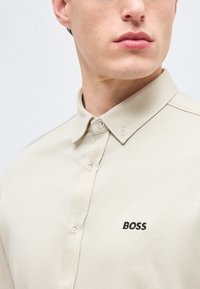 Gros plan d'une personne portant une chemise beige boutonnée avec un col pointu et "BOSS" brodé en noir sur la poitrine.