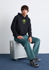 Juodas džemperis su žaliu grafikų, žydri kelnės ir juodai pilki sportbačiai. Sėdi ant sidabrinės tekstūruotos saugojimo dėžės su ratukais.