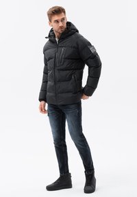 Schwarze Pufferjacke mit Kapuze, ausgestattet mit Reißverschlusstaschen, gerippten Bündchen und einem gesteppten Design. Kombiniert mit dunklen, verwaschenen Jeans und schwarzen Stiefeln.