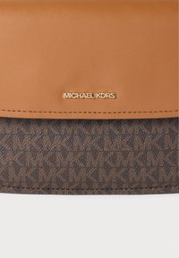 Καφέ δερμάτινο πορτοφόλι clutch με υφασμάτινο σχέδιο και χρυσά μεταλλικά στοιχεία. Διαθέτει το λογότυπο «MICHAEL KORS» εμφανώς τοποθετημένο. Σχεδιασμός επίπεδος και ορθογώνιος.