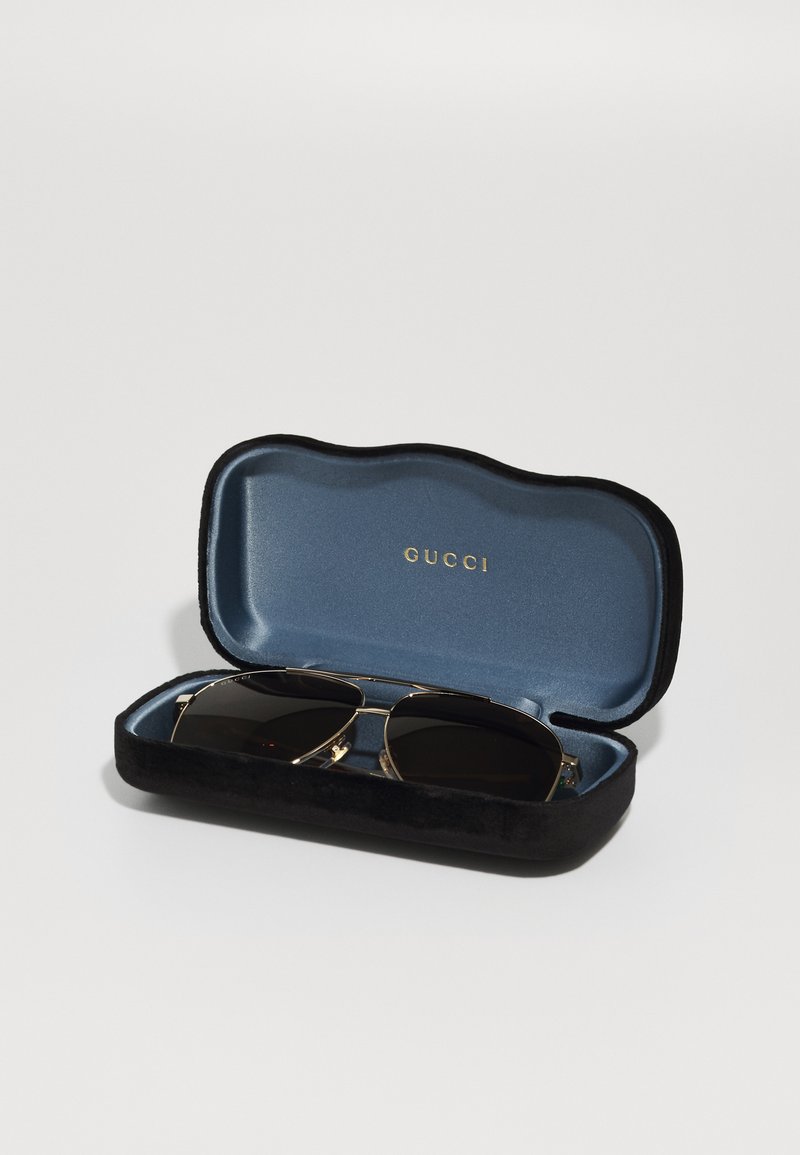 Custodia per occhiali da sole in velluto nero con interno blu, contenente occhiali da sole con lenti scure e montatura in metallo dorato. Logo stampato in oro all'interno.