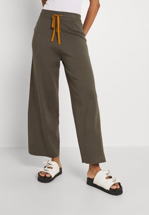 Personne portant un pantalon large olive avec un cordon orange et des sandales blanches à enfiler avec des semelles épaisses noires.