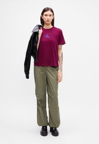 Βουργουνδής βαμβακερή μπλούζα με μπλε λογότυπο 'CK', συνδυασμένη με ελαιοπράσινα παντελόνια φαρδιάς γραμμής και μαύρες πλατφόρμες; το μοντέλο κρατά μια μαύρη δερμάτινη ζακέτα.