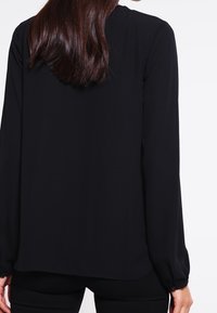 Black long-sleeve blouse with a smooth texture, featuring a round neckline and elastic cuffs, tapering at the hem for a slightly relaxed fit.