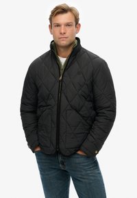 Chaqueta negra acolchada con cuello alto, dos bolsillos frontales y cierre de cremallera. Llevada sobre un suéter verde, combinada con jeans azules.