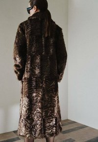 Cappotto di pelliccia sintetica marrone con un motivo testurizzato, maniche lunghe e colletto alto, che arriva all'altezza del polpaccio, indossato con occhiali da sole.