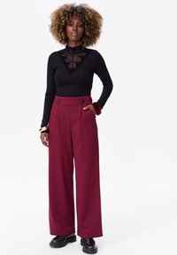 Zwarte lange mouwen top met stippen kantdetail, gecombineerd met burgundy wijde broek met zijzakken en knoopaccenten. Zwart schoeisel.