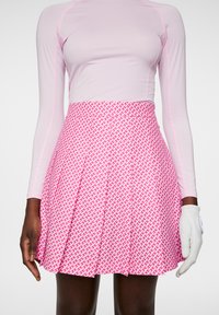 Vaaleanpunainen, laskostettu hame, jossa on geometrinen valkoinen kuvio, yhdistettyään tiukkaan pitkähihnaiseen puseroon. Valkoinen golfkäsine näkyy osittain.