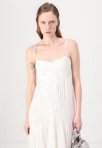 Rochie albă plisată cu bretele subțiri, bust texturat și fustă vaporoasă. Modelul ține o geantă gri cu un design strâns.