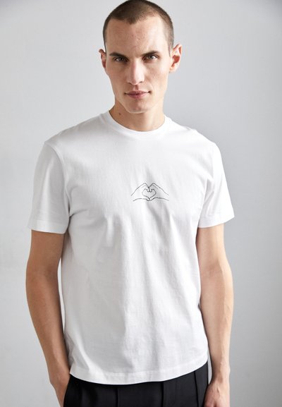 Camiseta de algodón blanca con un dibujo minimalista en línea negra de dos manos formando un corazón en el pecho. Manga corta, cuello redondo.