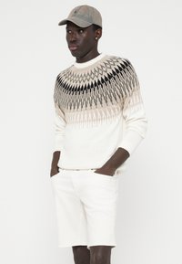 Geometrikus mintákkal díszített kötött pulóver krém, bézs és fekete színekben; fehér sorttal viselve. A krém színű baseball sapka teszi teljessé az outfitet.