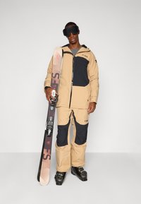 Beige og svart skikostyme med hette, slitesterkt stoff og glidelåslommer, båret av en person som holder ski med bindinger, svarte skibriller på.