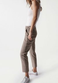 Femme se tenant dans un pantalon carreauté beige, un débardeur blanc et des baskets blanches sur fond blanc, de profil.