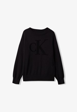Bambino con capelli castano chiaro in piedi con le mani in tasca indossa un maglione nero CK, jeans grigi sbiaditi e sneakers bianche su sfondo grigio.