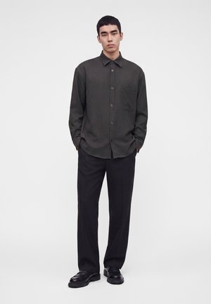 Homme debout les mains dans les poches, portant une chemise sombre à boutons, un pantalon noir et des chaussures noires sur un fond blanc uni.