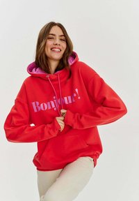 Červená mikina s kapucňou a ružovými akcentmi, s nápisom "Bonjour!" v hravom písme. Má predné vrecko a šnúrku na kapucni.