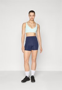 Vaaleansininen urheiluliivi ja tummansiniset korkeavyötäröiset shortsit, molemmat joustavaa kangasta. Mallilla on mustat lenkkarit ja valkoiset sukat.