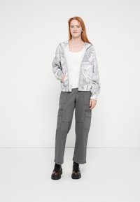 Pantalones cargo grises combinados con una camiseta blanca y una chaqueta bomber gris clara con estampado. La chaqueta tiene capucha y puños elásticos. Botas negras completan el conjunto.