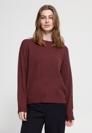 Jeune femme aux cheveux bruns portant un pull bordeaux à manches longues et un pantalon noir, debout devant un fond blanc uni.