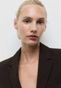 Hnědý blazer s vroubkovaným klopem, jedním knoflíkem a texturovanou látkou. Modelka má rovné blond vlasy a minimální makeup.