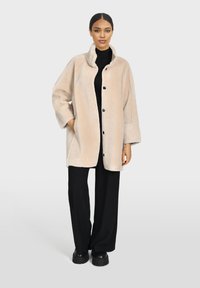 Abrigo beige, fuzzy y de gran tamaño con un cuello alto y botones negros, llevado sobre un suéter negro de cuello alto y pantalones negros de pierna ancha.