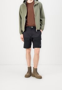 Olivgrön huva jacka, brun t-shirt, svarta cargo shorts med stora fickor och bruna snörstövlar. Texturerade material, funktionell design.