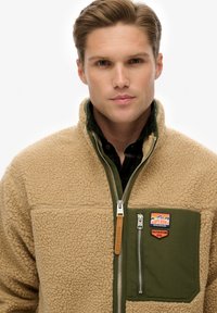 Beige fleecejack met een groen nylon borstzakje, ritssluiting, leren ritstrekker en merkpatches op de linkerborst. Gestructureerde stof.