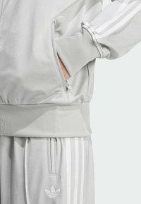 Světle šedý teplákový komplet s žebrovanými manžetami, zipovými kapsami a třemi bílými pruhy na rukávech a odpovídajících kalhotách. Jemný detail loga na lemu.