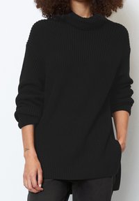 Maglione lavorato a maglia nero con colletto alto, texture a coste e maniche arrotolate. Presenta uno spacco laterale per maggiore comodità e una vestibilità rilassata.