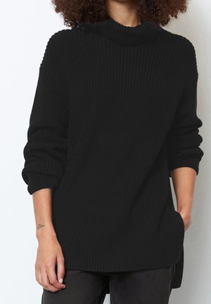 Pull en tricot noir avec un col haut, texture côtelée et manches retroussées. Il dispose d'une fente sur le côté pour plus de confort et d'une coupe décontractée.