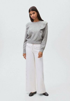 Vrouw met lang donker haar, gekleed in een grijze gerimpelde trui, wijde witte broek met zijkant studs en zwarte gevlekte slippers, tegen een effen achtergrond.