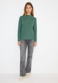 Pull en tricot côtelé vert avec un col montant, associé à un jean évasé gris et des baskets beiges. Le modèle pose devant un fond blanc.