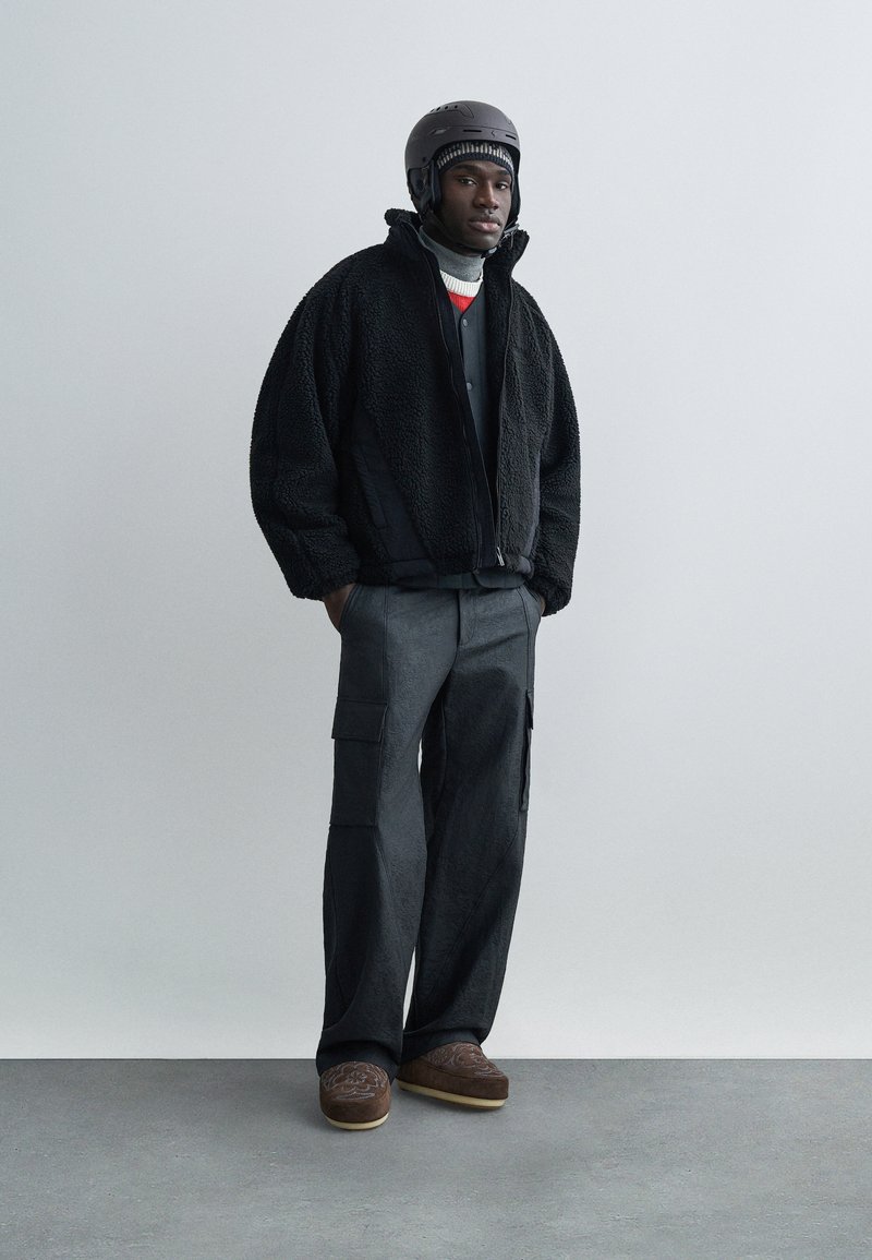 Man wearing black helmet, black fleece jacket, gray cargo pants, patterned brown shoes, and layered knitwear standing against plain gray wall.