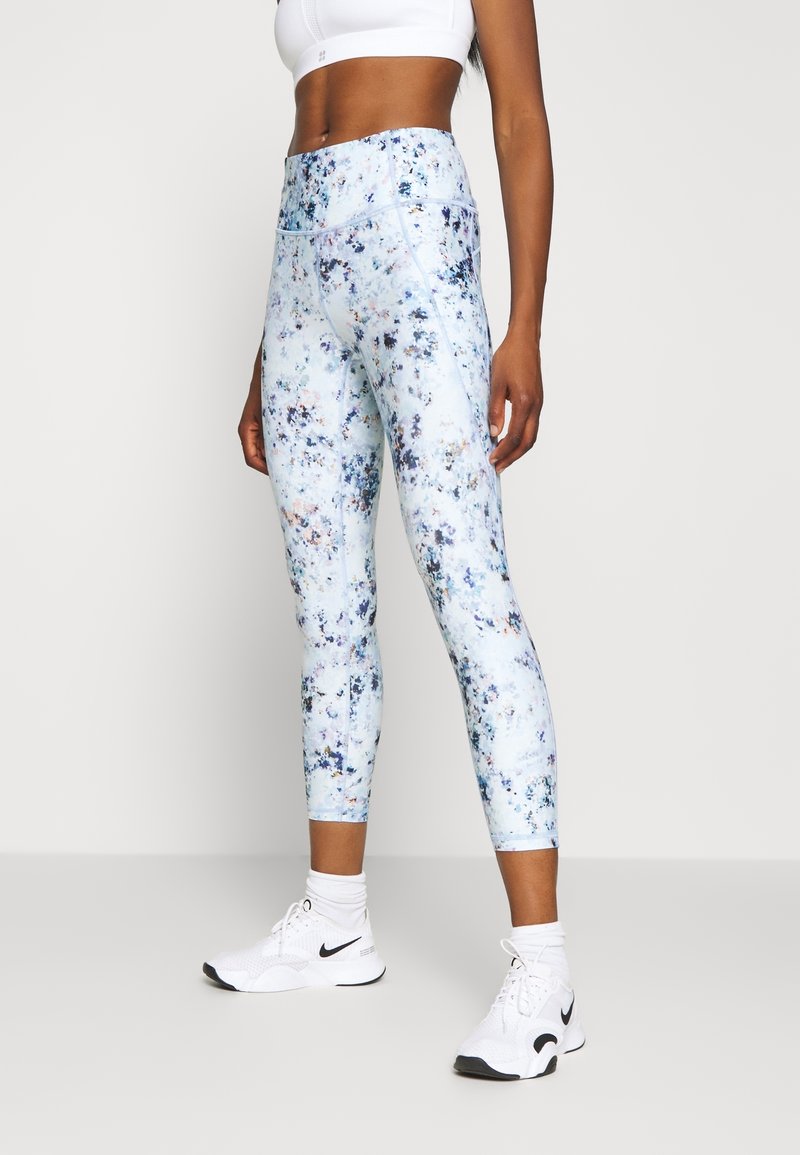 Femme portant un top de sport blanc court, des leggings taille haute bleu et blanc mouchetés, des chaussettes blanches et des baskets blanches Nike, debout.