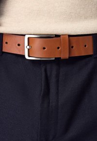 Ceinture en cuir marron avec une boucle rectangulaire en argent, comprenant plusieurs trous pour les réglages. Portée sur un pantalon en tissu foncé.
