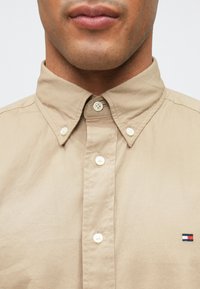 Camicia beige con bottoni e colletto button-down indossata da una persona, caratterizzata da un piccolo logo rettangolare rosso, bianco e blu sul petto.