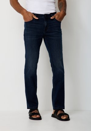 Man wearing dark blue jeans, white tank top, black sandals, standing with hands in pockets against a plain white background.