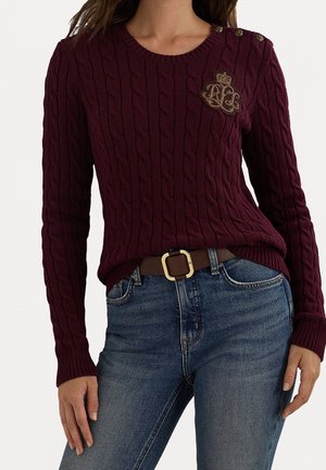 Femme portant un pull en maille torsadée bordeaux avec des boutons dorés sur une épaule et un blason brodé, associé à un jean bleu et une ceinture marron.