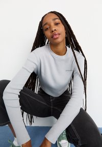 Šviesiai pilkas įribbintas ilgomis rankovėmis marškinėliai su apvaliu iškerpimu. Sujungti su juodomis džinsinėmis kelnėmis, aukštu juosmeniu. Sportbačiai turi žalias detales.