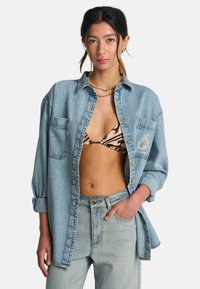 Jasnoniebieska koszula z denimu o luźnym kroju, z dwoma kieszeniami z przodu i detalem z logo. Noszona na wzorzysty bikini i dobrana do jeansów w tym samym kolorze.