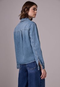 Helles blaues Denim-Hemd mit langen Ärmeln und seitlichen Schlitzen, ausgestattet mit einem klassischen Kragen und einem lässigen Schnitt, getragen mit dunkelblauen weit geschnittenen Jeans.
