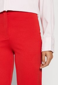 Pantalon rouge taille haute en tissu texturé, associé à une chemise à boutons rose clair. Poignet visible avec couture lisse au niveau du poignet.