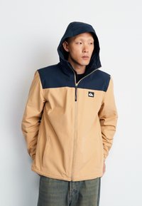 Beige hooded jacket with dark navy shoulder panels, zip-up front, and logo patch. Smooth texture and relaxed fit suitable for casual wear.