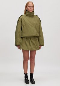 Groene, oversized jas met een hoge kraag en dubbele knopen, gedragen met een bijpassende gathered minirok, gecombineerd met zwarte sokken en schoenen.