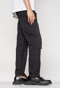 Pantalones cargo negros con detalles desgastados, con un bolsillo lateral, combinados con zapatos negros y una camisa blanca encima.