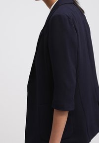 Námořnický blejzr se třičtvrtinovými rukávy, vyrobený z texturované tkaniny, s klopovým límcem a jednou přední kapsou na levé straně.