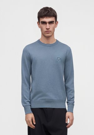 Jeune homme portant un pull léger bleu clair à manches longues et un pantalon noir, debout devant un fond blanc uni.