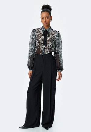 Blusa negra transparente con estampado floral, mangas largas abullonadas y una corbata al cuello, combinada con pantalones de pierna ancha negros de cintura alta y zapatos de punta.
