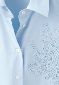 Cămașă albastru deschis cu guler ascuțit, cu nasturi albi și broderie florală detaliată într-o nuanță similară pe partea stângă.